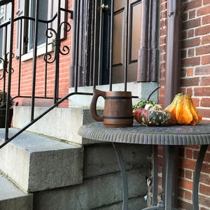 May include: A wooden beer mug with a dark brown finish and a black band around the middle sits on a grey metal table with three gourds. The gourds are orange, green, and black and white.