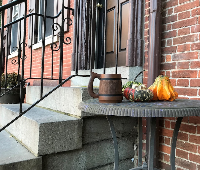 May include: A wooden beer mug with a dark brown finish and a black band around the middle sits on a grey metal table with three gourds. The gourds are orange, green, and black and white.
