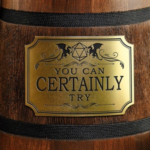 May include: A gold-colored metal sign with a black background and a black and white illustration of two dragons flanking a 20-sided die. The sign reads "You Can Certainly Try".