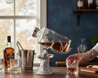 May include: A silver deer head decanter holder with a clear glass decanter filled with amber liquid. A person is holding a glass of amber liquid. Other barware items include a bottle of liquor, ice bucket, and glasses.