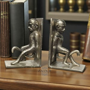 May include: A pair of silver-colored monkey bookends. Each bookend features a detailed monkey figure seated with its tail curled. The bookends are made of metal and are designed to hold books upright on a shelf or desk. The base of the bookends has the text "ZAIN R CREATION".