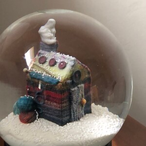 May include: A glass snow globe with a knitted house inside. The house is decorated with buttons and yarn. The snow globe is sitting on a black base.