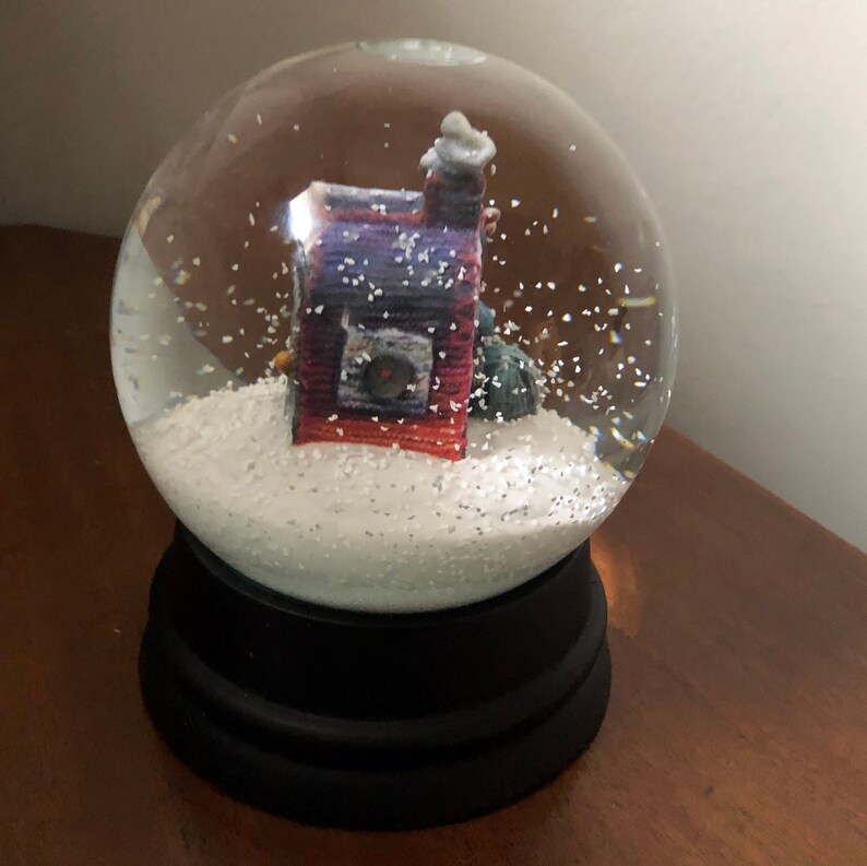 May include: A glass snow globe with a black base. Inside the globe is a small, red and purple house with a white chimney. The house is surrounded by white glitter.