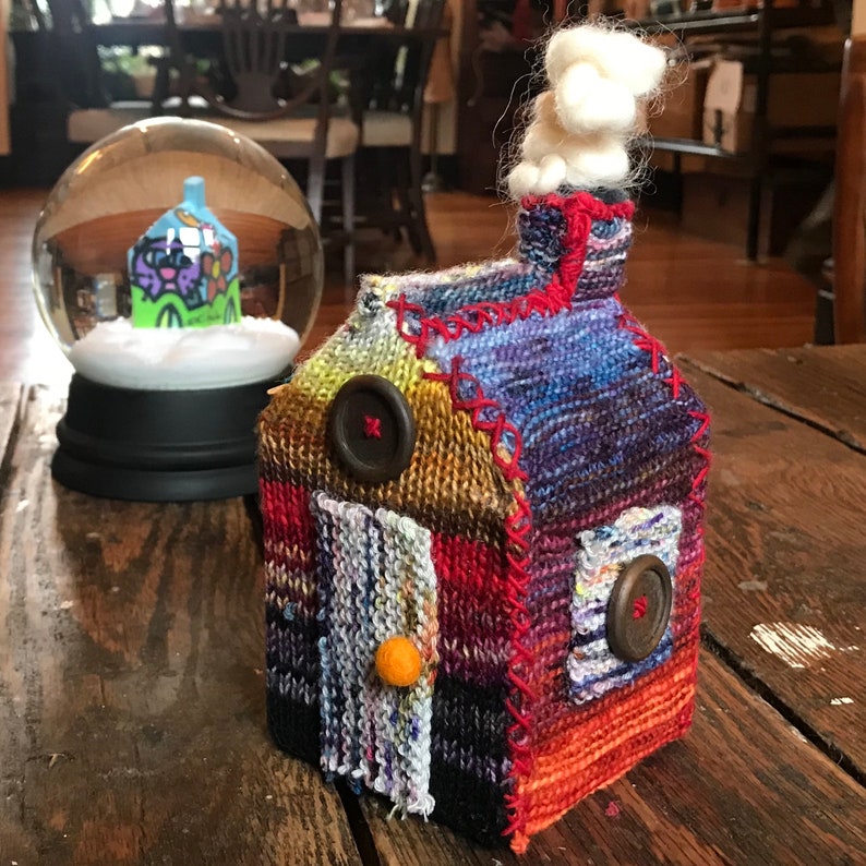 May include: A small knitted house with a white fluffy chimney, red trim, and colorful patchwork details. The house has a door with a yellow button and two brown buttons on the front.