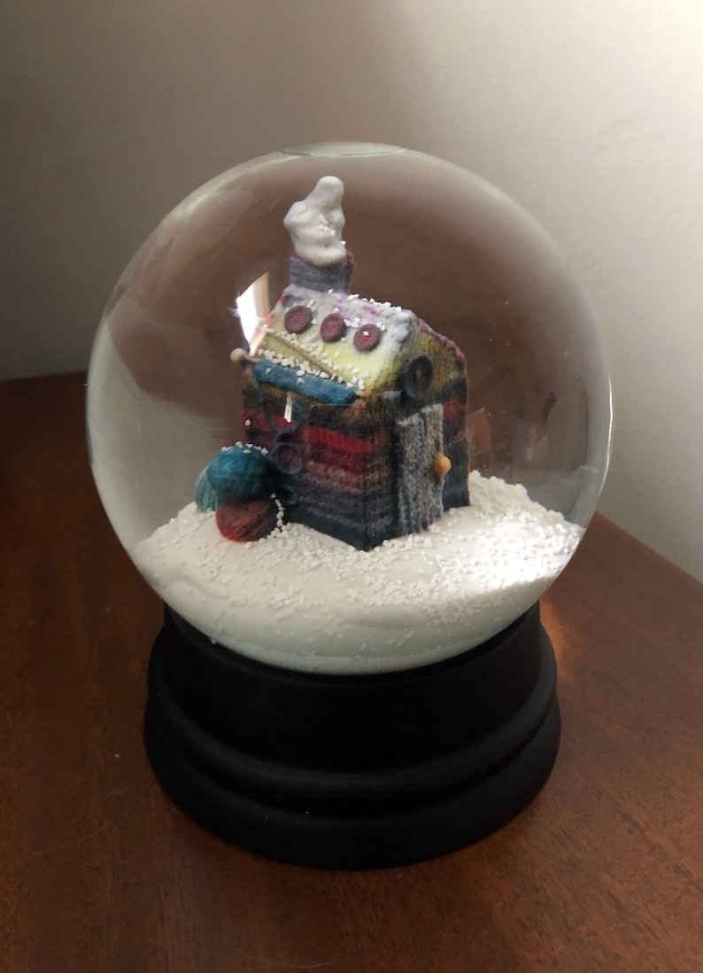 May include: A glass snow globe with a knitted house inside. The house is decorated with buttons and yarn. The snow globe is sitting on a black base.