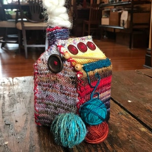 May include: A knitted house-shaped ornament with a white yarn chimney, red buttons, and yarn balls in shades of blue, red, and yellow.