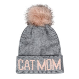 Puede incluir: Gorro de punto gris con un pomp&oacute;n de piel sint&eacute;tica rosa y las palabras "CAT MOM" bordadas en rosa en el pu&ntilde;o.