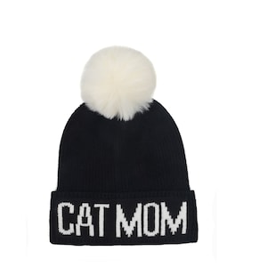 Puede incluir: Gorro de punto negro con un pomp&oacute;n blanco y las palabras "CAT MOM" bordadas en blanco en el pu&ntilde;o.