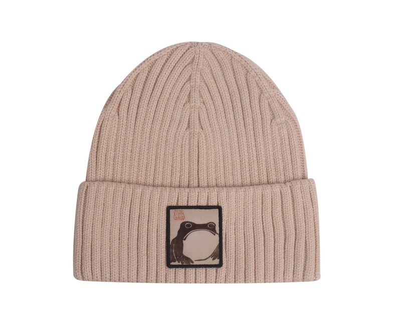 May include: A beige knitted beanie with a brown frog graphic and the text "The Frog King" on a white background.