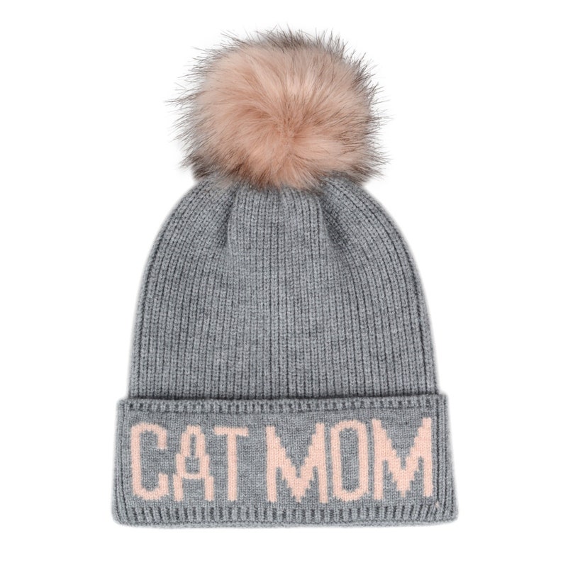 Puede incluir: Gorro de punto gris con un pomp&oacute;n de piel sint&eacute;tica rosa y las palabras "CAT MOM" bordadas en rosa en el pu&ntilde;o.