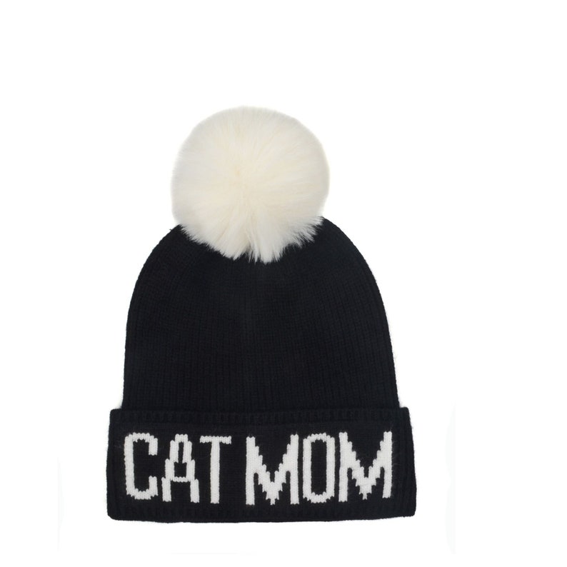 Puede incluir: Gorro de punto negro con un pomp&oacute;n blanco y las palabras "CAT MOM" bordadas en blanco en el pu&ntilde;o.