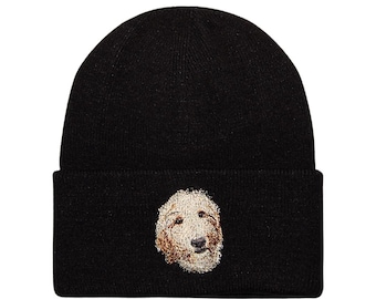 May include: Black knit beanie featuring an embroidered image of a dog's face. The dog's face is light brown and cream-colored, with dark brown eyes and nose. The beanie has a folded cuff.