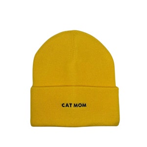 Puede incluir: Un gorro de punto amarillo con el texto "CAT MOM" bordado en negro.