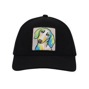 Op de afbeelding: Zwarte truckercap met een kleurrijk cartoon-dackel-patch op de voorkant.