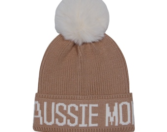 May include: A tan knit beanie with a white faux fur pom-pom on top. The hat features the text "AUSSIE MO" in white block letters across the brim. The beanie is a winter accessory.