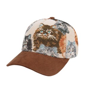 May include: A baseball cap with a brown suede brim and a cream-colored crown. The crown features a repeating pattern of various cat faces in shades of brown, gray, and blue. The cap has a brown button on top.