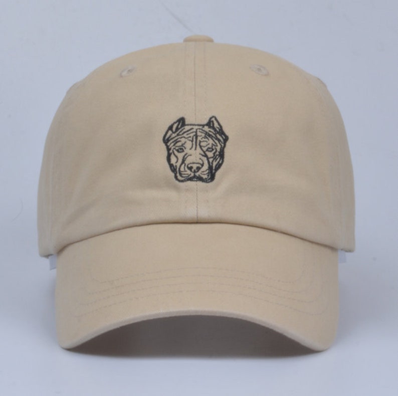 May include: A beige baseball cap with a black embroidered image of a pit bull's head.