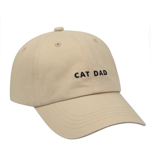 May include: A beige baseball cap with black embroidery that reads "CAT DAD".