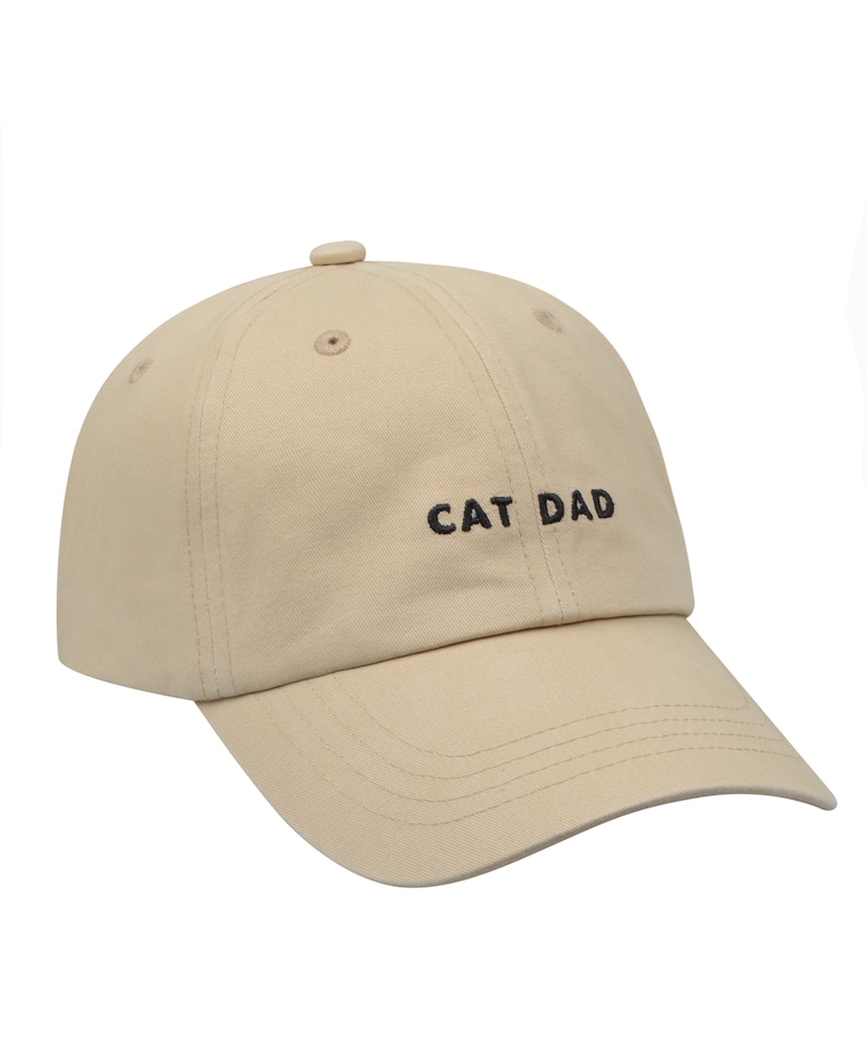 May include: A beige baseball cap with black embroidery that reads "CAT DAD".