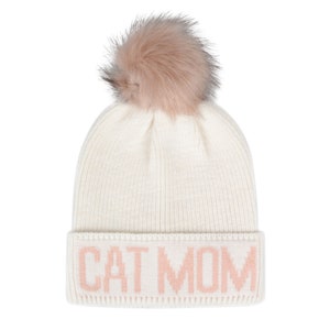 Puede incluir: Gorro de punto blanco con un pomp&oacute;n de piel sint&eacute;tica rosa y la frase "CAT MOM" en letras rosas.