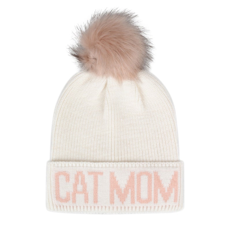 Puede incluir: Gorro de punto blanco con un pomp&oacute;n de piel sint&eacute;tica rosa y la frase "CAT MOM" en letras rosas.