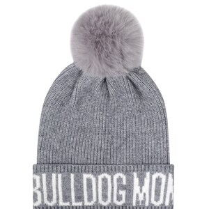 Pode incluir: Um gorro cinza de malha com um grande e fofo pompom cinza no topo. A aba dobrada apresenta o texto branco "BULLDOG MON". Um chap&eacute;u de inverno moderno para os meses mais frios.