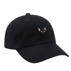 K&ouml;nnte beinhalten: Eine schwarze Baseballkappe mit einem gestickten schwarzen Katzenkopf mit gelben Augen.