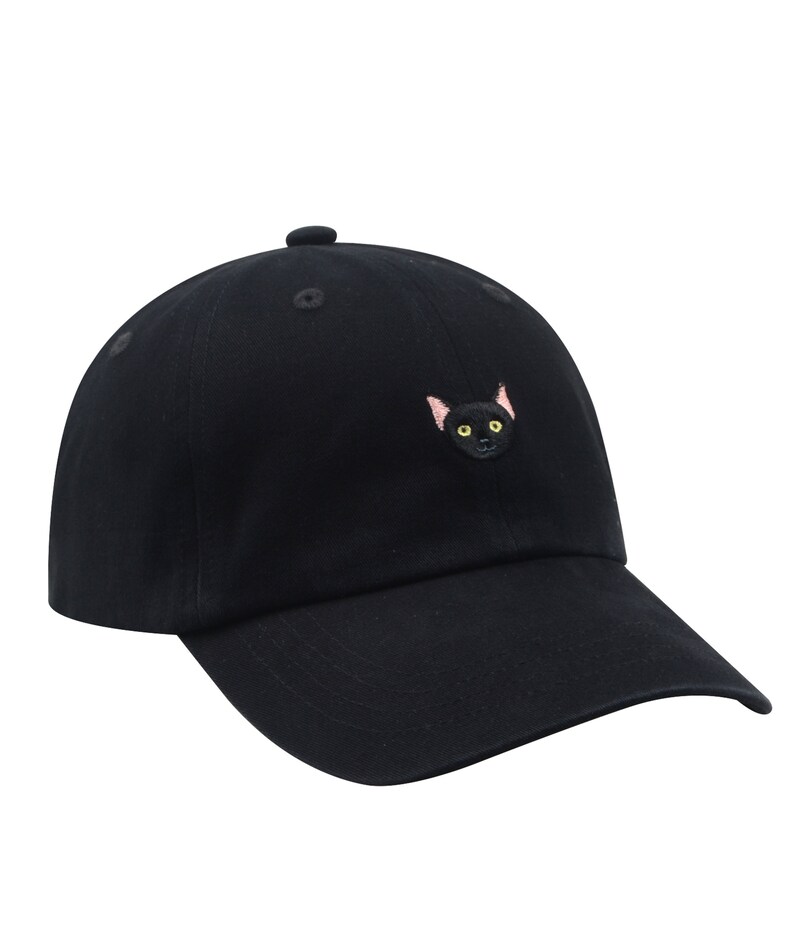 K&ouml;nnte beinhalten: Eine schwarze Baseballkappe mit einem gestickten schwarzen Katzenkopf mit gelben Augen.