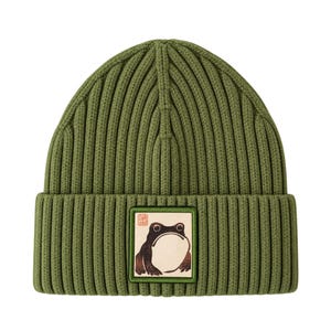 May include: An olive green ribbed knit beanie featuring a square patch on the front cuff. The patch displays a graphic illustration of a frog in a woodblock print style with a small red square seal mark in the upper left corner of the patch.