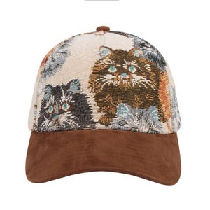 May include: A baseball cap with a brown suede brim and a patterned crown featuring multiple cat faces in shades of brown, gray, and cream. The cats have blue and green eyes. The cap has a brown button on top.