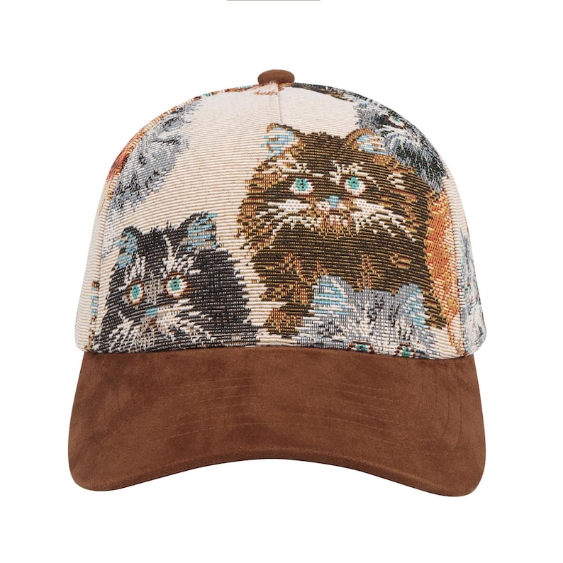 May include: A baseball cap with a brown suede brim and a patterned crown featuring multiple cat faces in shades of brown, gray, and cream. The cats have blue and green eyes. The cap has a brown button on top.