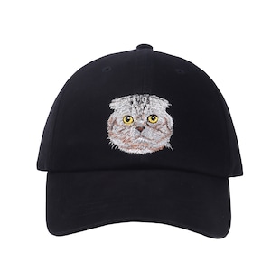 Peut inclure: Une casquette de baseball noire avec une image brodée d'un chat gris et blanc aux yeux jaunes.
