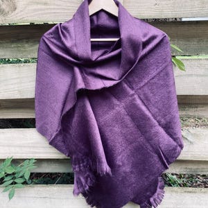 May include: A deep purple, fringed shawl draped over a wooden hanger. The shawl is made of a soft, luxurious fabric and has a subtle sheen.