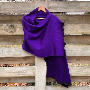 May include: A vibrant purple shawl with black fringe hangs on a wooden hanger. The shawl is draped over a wooden fence, showcasing its rich color and texture. The fringe adds a decorative element to the edges of the shawl.