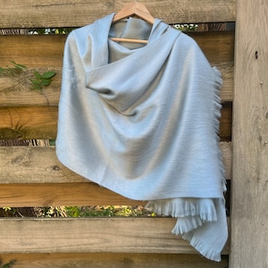 May include: A light gray shawl with a soft, textured appearance, displayed on a wooden hanger. The shawl has a draped design with fringed edges, creating a flowing and elegant look. The background is a wooden fence.