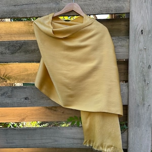 May include: A golden-yellow shawl with a fringed edge hangs on a wooden hanger. The soft, woven fabric drapes elegantly, showcasing its texture. The shawl is displayed against a rustic wooden fence.