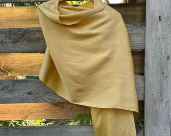 Puede incluir: Un chal de color amarillo dorado con un borde con flecos cuelga de una percha de madera. La suave tela tejida se drapea elegantemente, mostrando su textura. El chal se exhibe contra una valla de madera rústica.