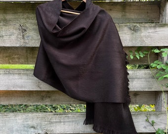 May include: A dark brown shawl with a fringed edge hangs on a wooden hanger against a weathered wooden fence. The shawl is draped to show its texture and shape, with a soft, flowing appearance.
