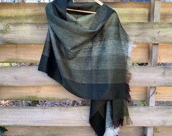 May include: A dark green and gray shawl with a striped pattern, hanging on a wooden hanger. The shawl has a fringed edge and is draped over a wooden fence.