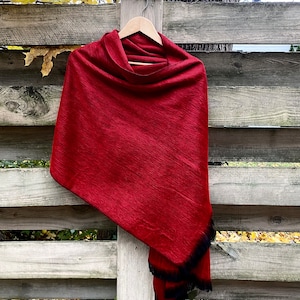 May include: A deep red shawl with a black fringe hangs on a wooden hanger against a weathered wooden fence. The shawl is draped to show its texture and shape, suggesting a soft, warm material.