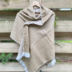May include: A beige shawl with a white fringe hanging on a wooden hanger. The shawl is made of a soft, textured fabric.