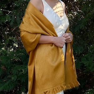 May include: A mustard yellow rectangular shawl featuring a fine, textured weave and a fringed hem along the bottom edge. The fabric has a subtle sheen and is draped over the shoulders, showcasing the length and the soft, flowing drape of the material.