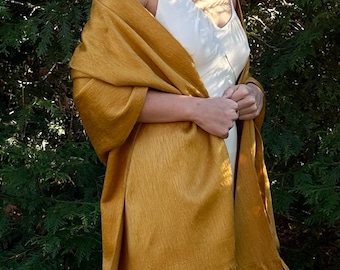 Puede incluir: Un chal rectangular de color amarillo mostaza con un tejido fino y texturizado y un dobladillo con flecos en el borde inferior. La tela tiene un brillo sutil y está drapeada sobre los hombros, mostrando la longitud y la caída fluida.