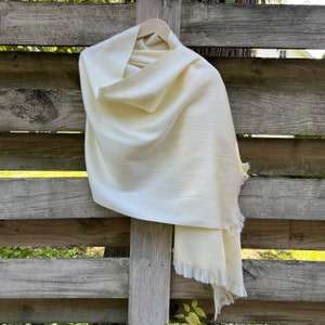 May include: A cream-colored shawl with a fringed edge hangs on a wooden hanger. The soft, woven fabric drapes over the hanger, showcasing its texture. The shawl is displayed against a weathered wooden fence.