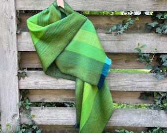 May include: A green and blue striped scarf is displayed on a wooden fence. The scarf is made of a soft material and has a gradient of green shades, with blue accents at the ends. The scarf is hanging from a wooden hanger.