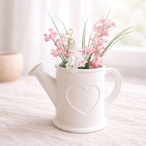 Op de afbeelding: Een witte keramische gieter-vormige vaas met een hartvormige uitsparing, gevuld met roze en witte bloemen en groene stelen. De vaas heeft een handvat en een tuit. De opstelling staat op een neutraal gekleurd oppervlak.