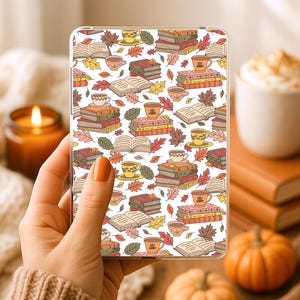 Peut inclure: Une coque de téléphone transparente avec un motif répété de livres, tasses à thé et feuilles d'automne dans des tons de marron, orange et jaune. La coque est tenue dans une main. L'arrière-plan comprend une bougie allumée, des citrouilles et une tasse de café.