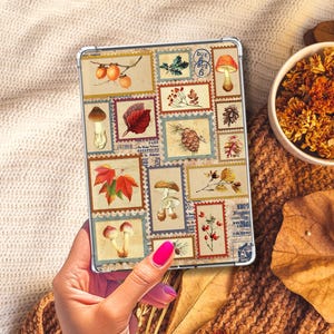 Könnte beinhalten: Eine Tablet-Hülle mit einer Collage aus Vintage-Briefmarken-Designs. Die Briefmarken zeigen botanische Illustrationen von Pilzen, Blättern und Beeren in herbstlichen Farben. Die Hülle hat einen silbernen Rahmen und wird von einer Hand gehalten.