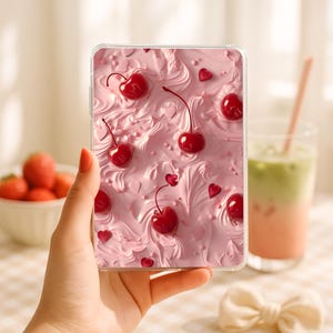 Peut inclure: Une coque de téléphone transparente avec un motif rose texturé, ornée de cerises rouges et de formes de cœur. L'étui est tenu dans une main, avec un arrière-plan flou comprenant une boisson et des fraises.