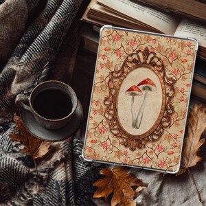 Könnte beinhalten: Eine Tablet-Hülle mit einer Vintage-Illustration von zwei rot gekappten Pilzen in einem ovalen Rahmen. Der Hintergrund hat ein beiges Blumenmuster mit Herbstblättern. Eine Tasse Kaffee und eine karierte Decke sind im Hintergrund.